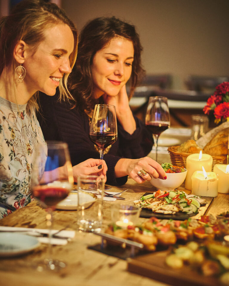 Two women look at a selection of food on a wooden table in front of them.
