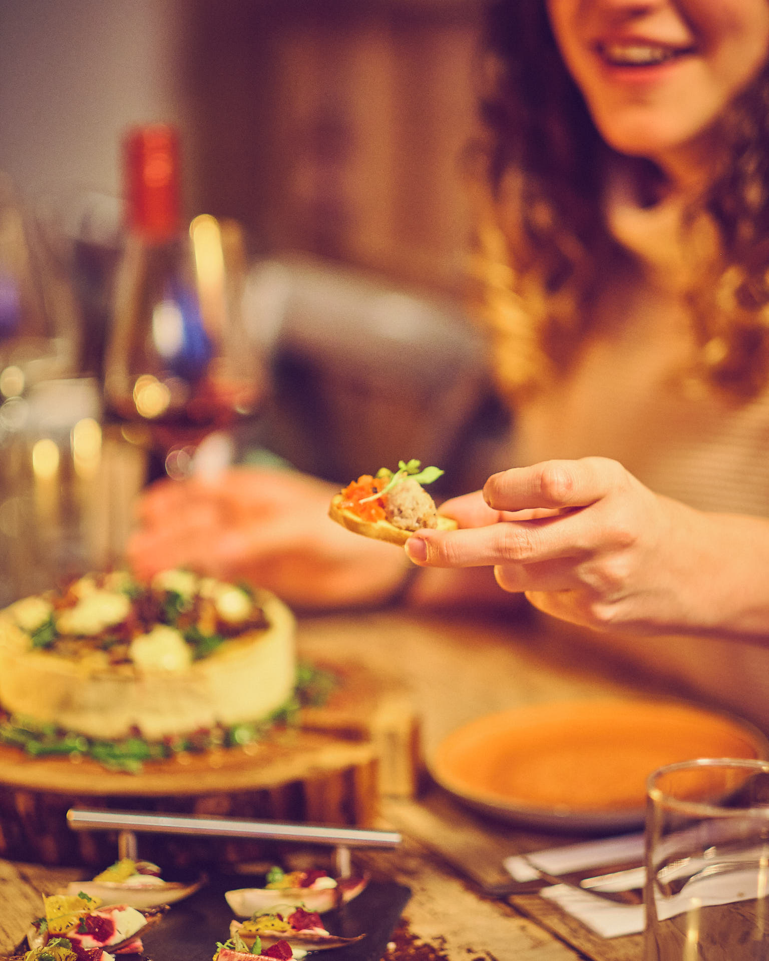 A lady holds a small bite of party food whilst sat a table drinking wine.